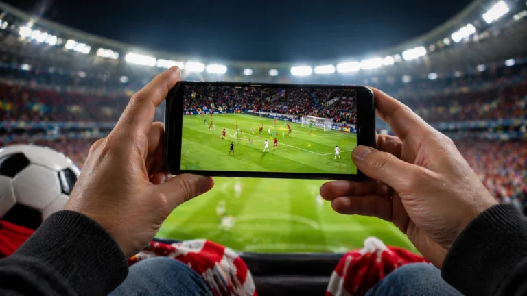 Pessoa segurando celular dentro de estádio lotado enquanto assiste partida de futebol ao vivo na tela, com bola e torcida ao fundo.